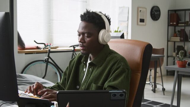 Medium side shot of focused young African American male programmer writing code for mobile app on desktop computer in startup office, listening to music, smartphone, tablet and laptop on desk