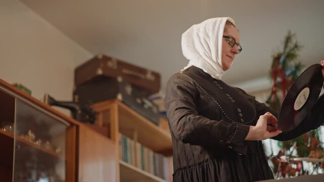 Woman in headscarf cleaning vinyl record, wearing glasses and acting as conservator in home studio, polishing grooves with soft cloth, inspecting under magnifier, shelves of albums and vintage