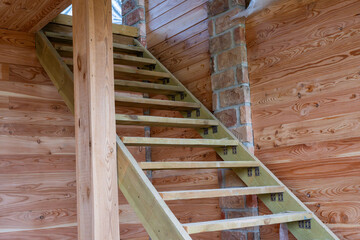A newly constructed wooden staircase with open risers stands inside an unfinished timber frame house or rustic log cabin. © Andrey