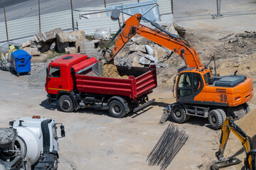 a bright orange wheeled excavator loading dirt and rubble into a red dump truck on an active, sunlit construction site. © Andrey