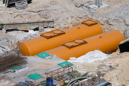 two large, bright orange cylindrical underground storage tanks resting in a dirt excavation pit during installation at an active construction site.