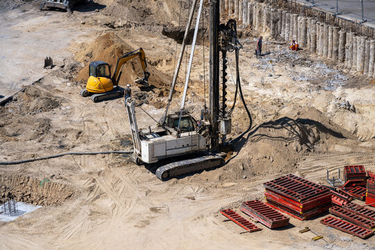 An active construction site featuring a heavy-duty crawler piling rig and a yellow mini-excavator performing earthworks and foundation preparation.