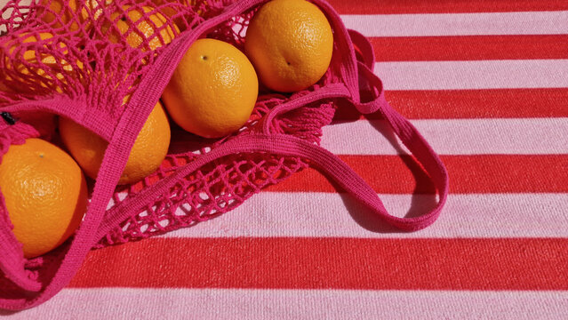 Fresh oranges spilling from a bright pink eco-friendly mesh shopping bag. A bold red and pink striped beach towel with natural sunlight casting distinct shadows. Space for text. 