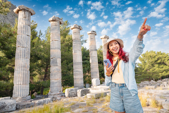 Happy woman tourist exploring ancient Greek ruins, standing by the Temple of Athena columns, holding a smartphone and pointing up