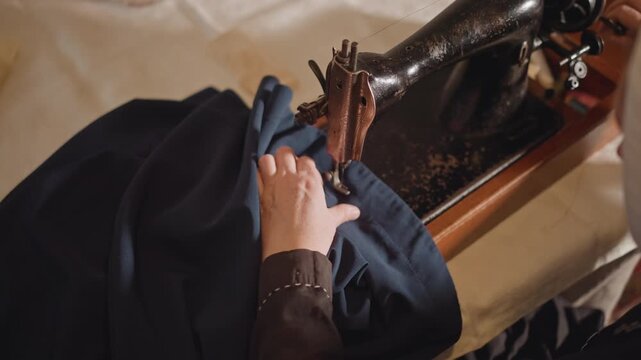 Skilled tailor meticulously working on deep blue textile at historic sewing machine. Closeup of focused tailor diligently hemming navy cloth using antique sewing device in cozy workshop environment