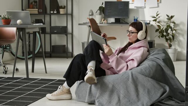 Full shot of young gen Z Caucasian female mobile application developer working on tablet while sitting comfortably in bean bag chair in startup office, using vibe coding assistant and enjoying music