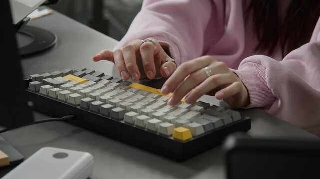 Close-up of hands of unrecognizable young female programmer typing code on computer keyboard, wearing rings on fingers and pink sweatshirt, while working on software on mobile application