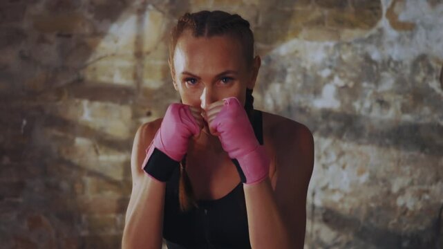 Young athletic woman with braided hair throwing punch wearing pink bandages during intense training session in gym with focused expression