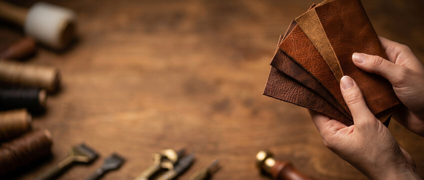 Close-up of artisan hands holding brown leather swatches over a rustic wooden workbench with blurred crafting tools. Ideal template for small business owners banner with copy space
