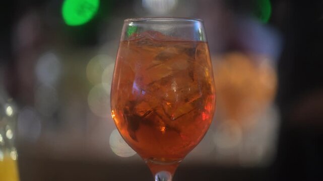 An aperol spritz drink on a glass at a lively bar during the evening with defocused background with a barman dancing