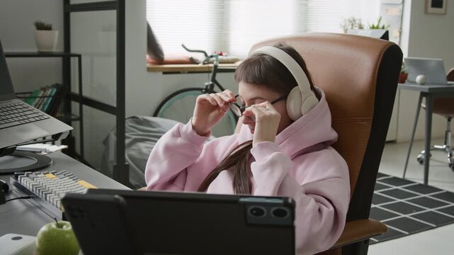 Medium side shot of tired Caucasian female web developer reclining in chair while working on code and testing on multiple computer devices in office, taking off glasses, rubbing face with exhaustion
