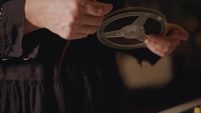 Closeup hands threading vintage film reel, careful fingers guide perforated strip onto spool, translucent frames catch warm light as projectionist prepares footage for playback and restoration.