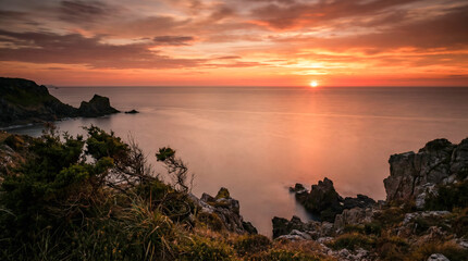 Fototapeta premium Puesta de sol desde un acantilado costero sobre un mar en calma en verano, con el agua pintada de naranja y rojo y larga exposición suavizando las olas.