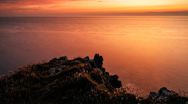 Puesta de sol desde un acantilado costero sobre un mar en calma en verano, con el agua pintada de naranja y rojo y larga exposici&oacute;n suavizando las olas.