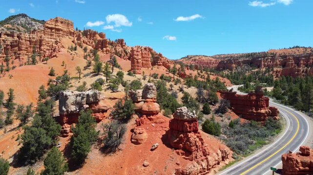 Smooth drone footage flying over a winding highway with cars amidst brilliant red rock spires and pinnacles in Dixie National Forest. Magical canyon landscape in Utah under a sunny blue sky.
