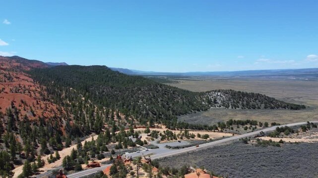 Smooth drone footage flying over a winding highway with cars amidst brilliant red rock spires and pinnacles in Dixie National Forest. Magical canyon landscape in Utah under a sunny blue sky.