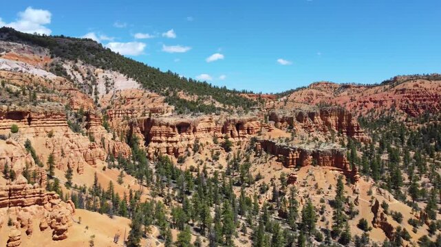 Smooth drone footage flying over a winding highway with cars amidst brilliant red rock spires and pinnacles in Dixie National Forest. Magical canyon landscape in Utah under a sunny blue sky.