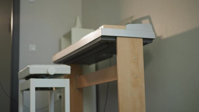 Digital piano with stool on carpet, professional coaching rehearsal in compact studio, wall light casting soft shadow on textured wall, tripod and music stand visible, coach preparing scores, focused