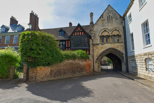 A historic stone archway and half-timbered building stand under a blue sky. A road leads through the arch, past a brick wal