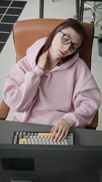 Medium vertical high-angle shot of young Caucasian female software developer in glasses massaging sore neck to relieve muscle tension while working on project on desktop computer in corporate office