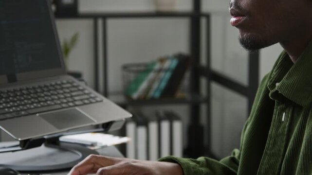 Tilting close-up of focused African American male programmer typing on computer keyboard, checking result on screen and smiling while vibe coding for mobile app in technology startup office