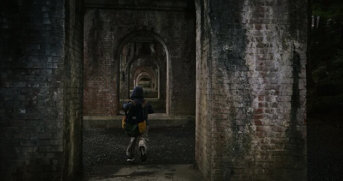 Boy with hoodie and backpack running through brick arches under Japanese bridge in slow motion