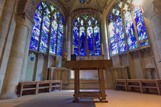 A serene Cathedral interior bathed in blue light from large, vibrant stained-glass windows. A simple wooden altar stands centrally at Gloucester Cathedral - Gloucester - Gloucestershire - UK
