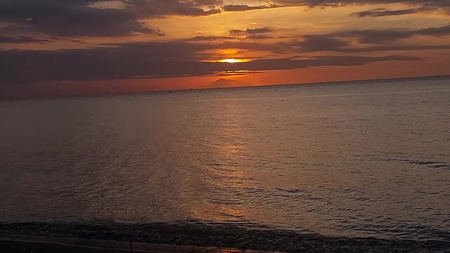 Beautiful golden sunset over a black sand beach with calm sea water and colorful evening sky. Peaceful coastal scenery with warm sunlight reflection, located at Singaraja, Bali, Indonesia.