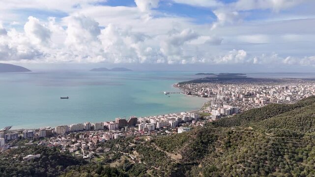 Wide aerial tracking shot flying forward over a dense coastal cityscape, turquoise sea waters, and a distinct brown sediment plume along the Bay of Vlor&euml; coastline in Albania