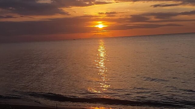 Beautiful golden sunset over a black sand beach with calm sea water and colorful evening sky. Peaceful coastal scenery with warm sunlight reflection, located at Singaraja, Bali, Indonesia.