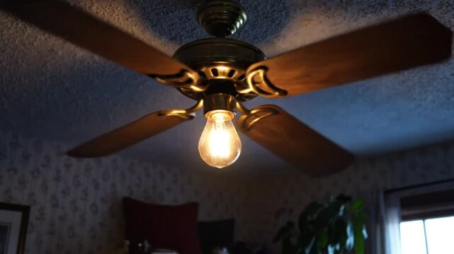 Low angle view of ceiling fan spinning with its incandescent light bulb glowing brightly in dim room
