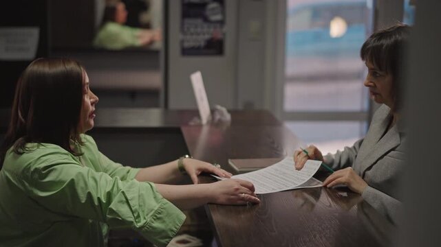 Front desk intake consultation at music school reception, student and coach reviewing paperwork across counter, papers and pen exchanged, framed certificates in background, calm professional mood