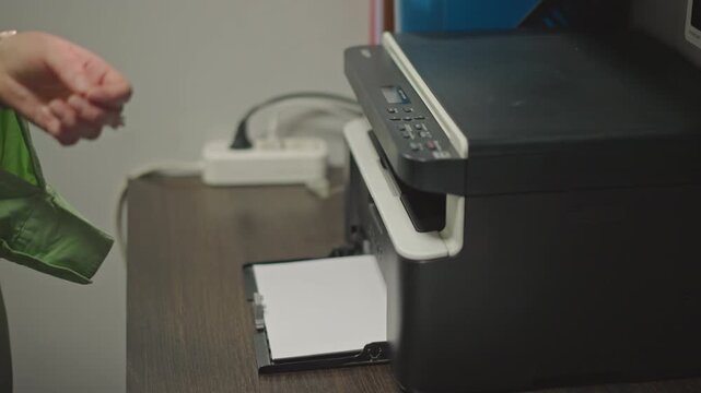 Music teacher prints sheet music while retrieving fresh pages from compact office printer, closeup on hand guiding paper tray, soft task lighting on tidy desk, visible cables and supplies, calm