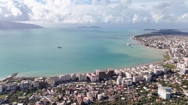Wide aerial tracking shot pulling back over dense coastal high-rises, urban streets, and the turquoise waters of the Bay of Vlor&euml; under a cloudy blue sky in Albania