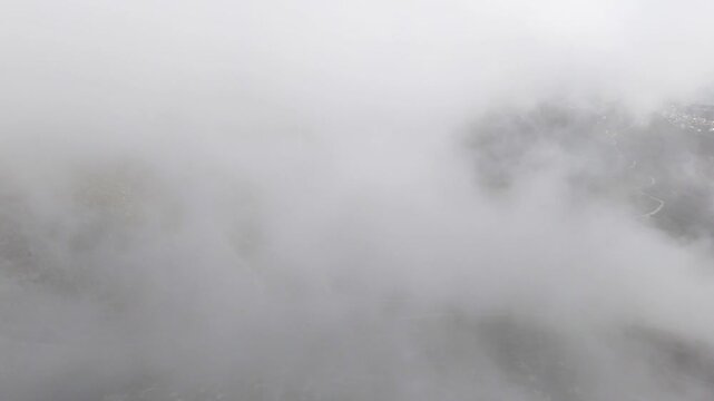 High-angle aerial shot moving forward directly through thick white fog and swirling clouds over the dramatic winding switchbacks of the Llogara Pass mountain road in Albania