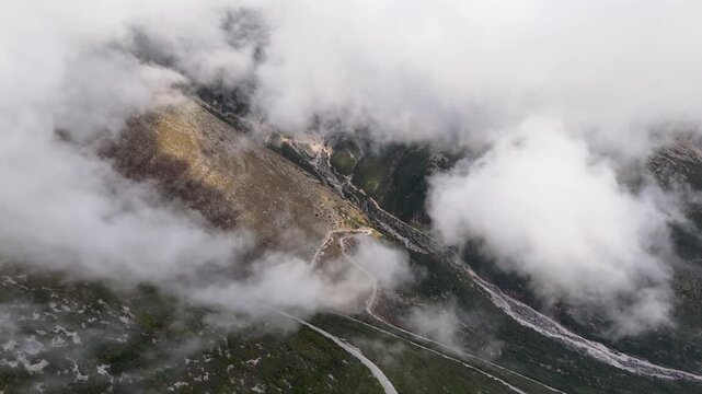 High-angle aerial shot rising upward through swirling white clouds, revealing steep rocky slopes and a winding serpentine mountain road below at Llogara Pass, Albania
