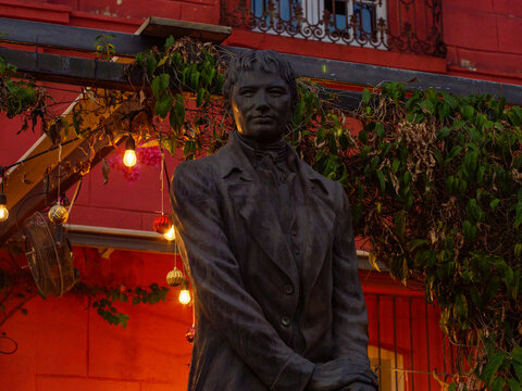 Estatua de bronce de personaje hist&oacute;rico frente a una colorida fachada roja con luces c&aacute;lidas y vegetaci&oacute;n