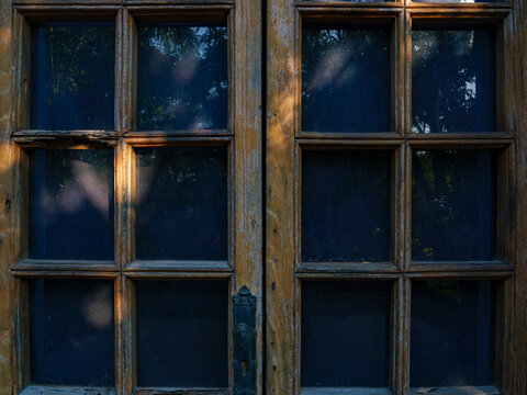 Detalle en primer plano de puertas r&uacute;sticas de madera antigua con cristales que reflejan &aacute;rboles bajo una luz c&aacute;lida