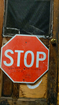 Vertical close-up of a weathered red stop sign featuring handwritten expletive graffiti about leaving a door open, mounted on a rustic, distressed wooden door with a screen window.