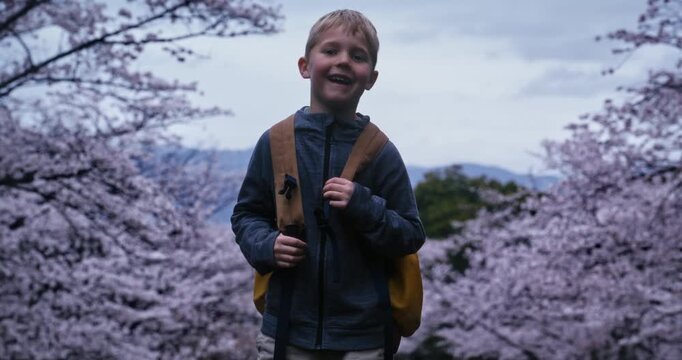 Happy boy with backpack turning around while standing among cherry blossom trees