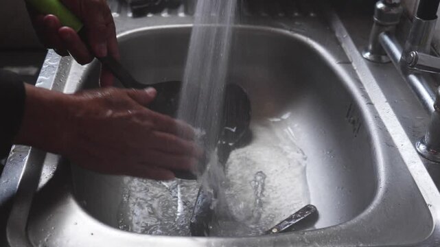 Person washing a saucepan in a stainless steel kitchen sink with running water from the faucet, performing daily housework