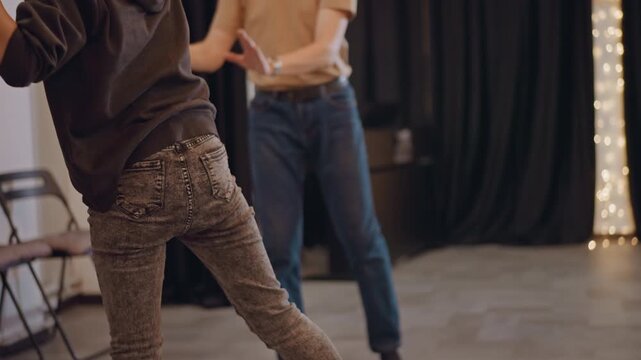 Close rehearsal white woman student practicing choreography in studio hooded redhead performer executes squat and step sequences guided by instructor, studio floor and curtain backdrop with string