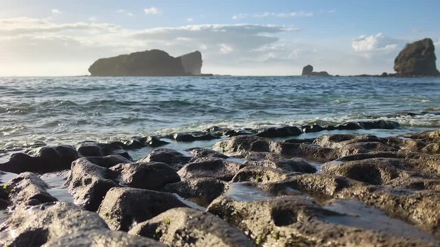 Slow motion shot of ocean waves gently crashing against unique volcanic rock formations on the beach, with a beautiful rocky island in the background. Papuma Beach, Jember, Indonesia.