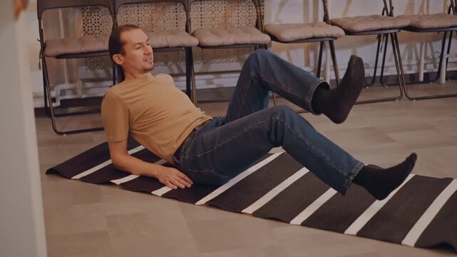 Musician teacher leg warmup on rug focusing on core mobility and breath casual instructor in jeans demonstrates alternating leg lifts on striped floor rug in classroom filled with stacked chairs, slow