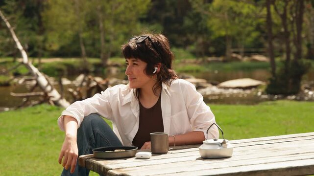 Keyframe 1: A woman sits at a picnic table. She wears a white shirt and jeans. Her expression is calm and relaxed. Sunlight filters through trees. Teapot and cup rest on the table