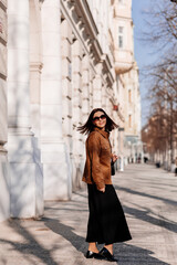 Obraz premium Beautiful young woman in sunglasses going for a walk in Prague city. Stylish girl in a brown jacket posing near a historic building facade on a sunny day. Urban fashion and lifestyle concept.