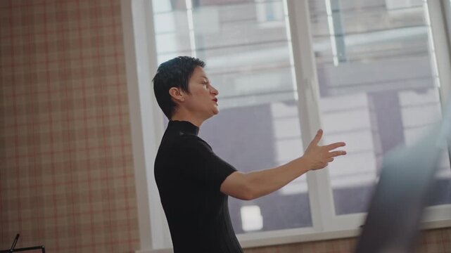 Asian vocal coach teaching in classroom, expressive hand gestures guiding breath and pitch during focused rehearsal, sunlit window and patterned wallpaper, black turtleneck, supportive instruction
