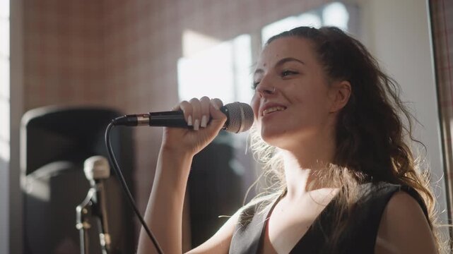 Singer smiling during vocal practice session, expressive phrasing and bright natural light, confident mic technique, upbeat mood with band equipment in background, musician delivering warm performance