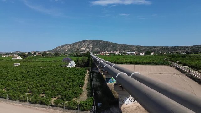 Aerial view of the large Tajo Segura water transfer pipeline crossing the fertile agricultural lands orchards and fields of Vega Baja under a clear blue sky towards a distant mountain