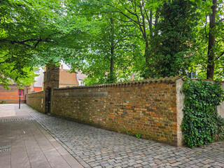 Fototapeta premium quiet pedestrian path bordered by a long brick wall and dense green trees. The scene suggests a residential or historic urban area with a calm atmosphere and natural surroundings
